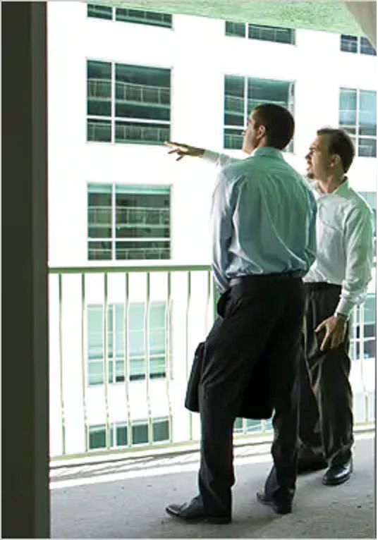 Two men in casual business attire stand on the exterior deck of a commercial building, one pointing to an adjacent property